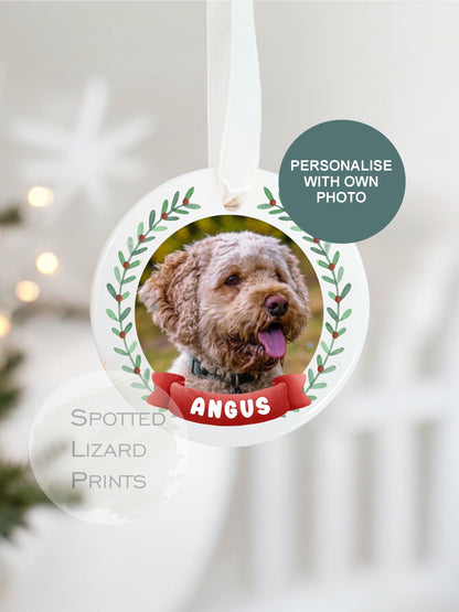 Personalised ceramic Christmas bauble featuring a photo of a dog, framed by a festive green wreath design and a red banner with the dogs name, hanging on a white ribbon