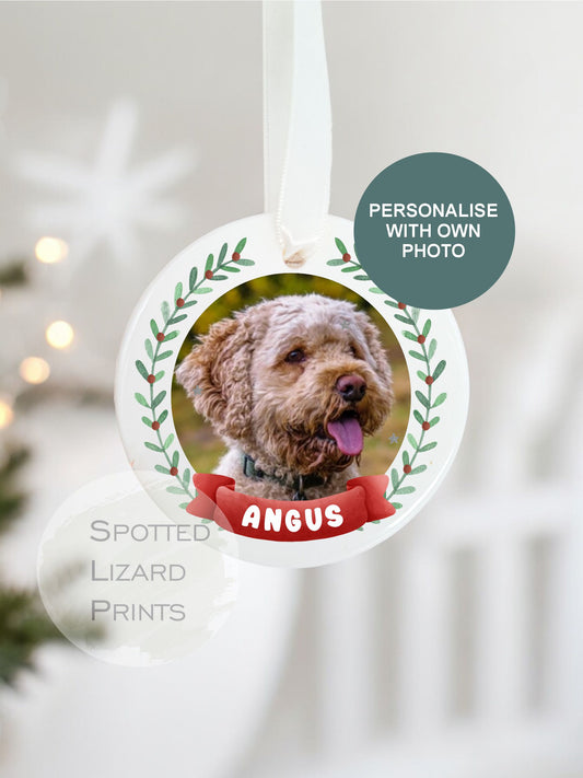 Personalised ceramic Christmas bauble featuring a photo of a dog, framed by a festive green wreath design and a red banner with the dogs name, hanging on a white ribbon